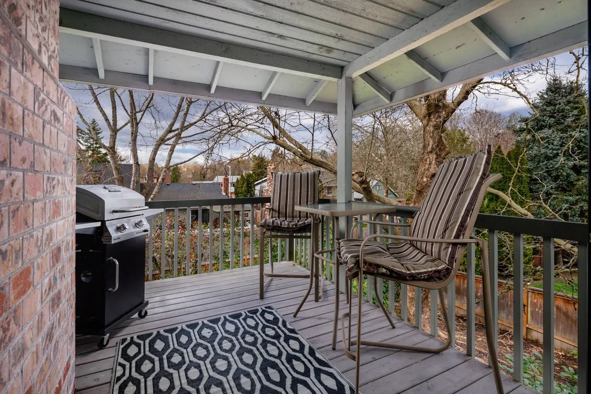 A covered outdoor deck featuring a modern grill, comfortable outdoor seating, an area rug, and a scenic view of treetops beyond the railing. The space is set up for barbecuing, relaxing, and dining, with a tranquil atmosphere perfect for enjoying nature in privacy.