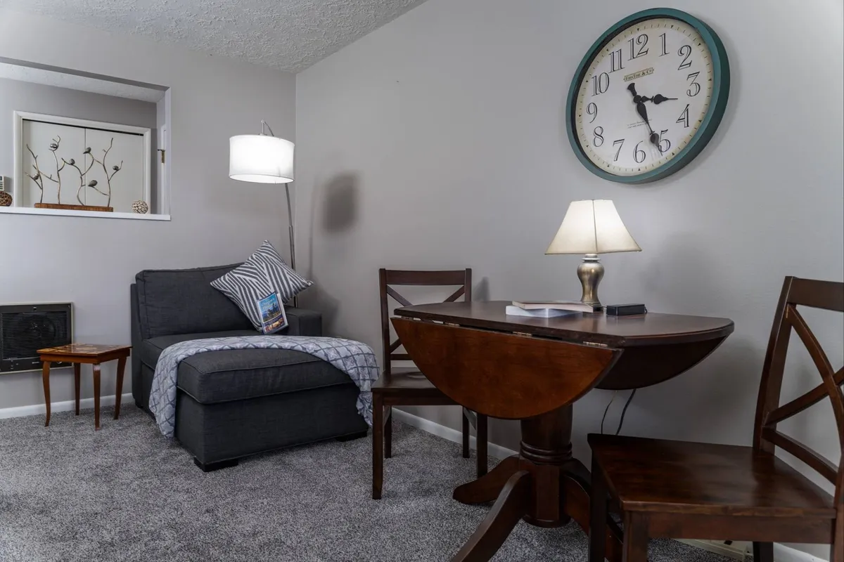 This image features a comfortable living space with a stylish sofa, a well-appointed dining table, a wall clock, and a lamp that casts a warm glow. The arrangement creates an inviting atmosphere, suggesting a perfect spot to relax after exploring the city or enjoy a quiet meal before heading out.