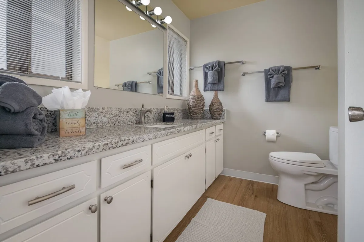 A modern, calming bathroom featuring a granite countertop, large mirror, towel rack, decorative vases, and neatly arranged towels. The space feels fresh and serene, ideal for morning routines or unwinding at night, with clear signs of thoughtful upgrades.