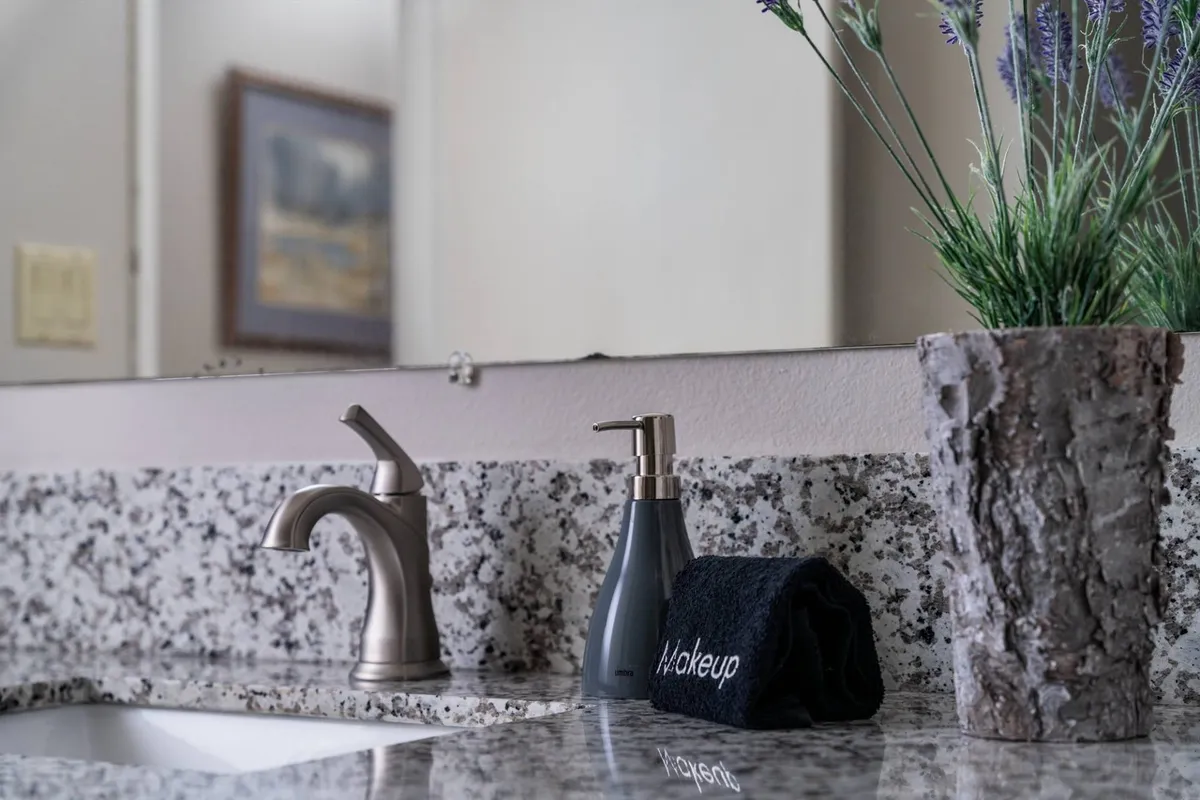 A granite bathroom countertop with a modern faucet, soap dispenser, plush towel, and a potted plant. Morning light pours in, enhancing the inviting and refreshing ambiance—ideal for relaxation and personal care.