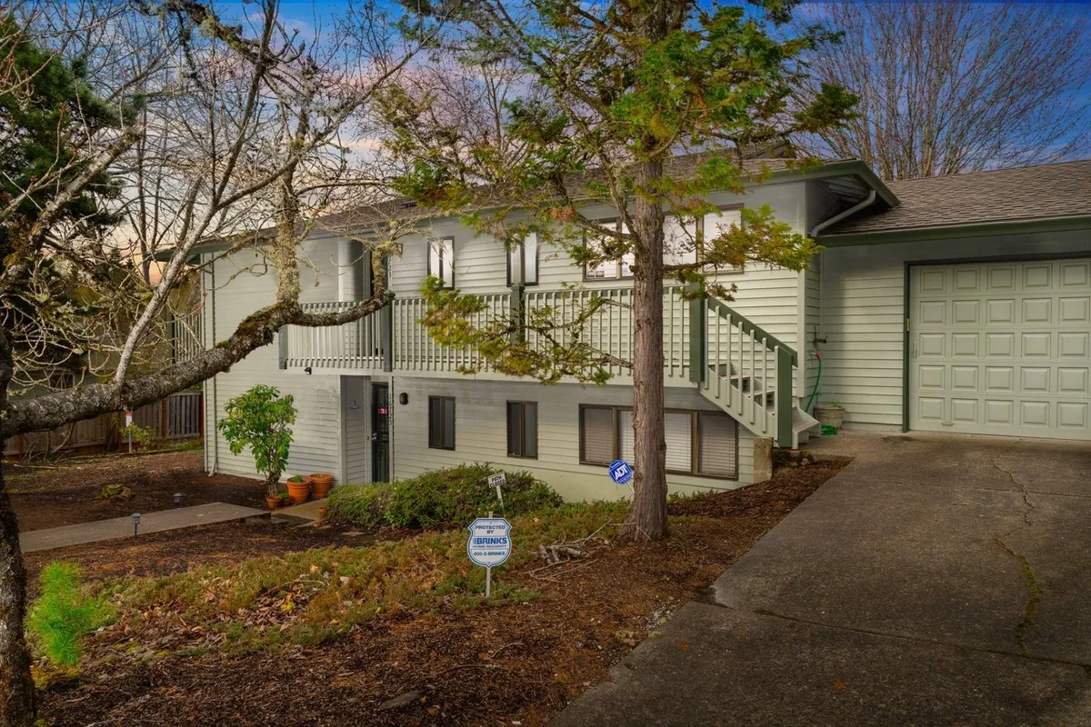 The image showcases the inviting exterior of Summercrest Cottage, featuring a welcoming front porch with cozy seating, a beautifully landscaped garden, and a sunlit driveway that accentuates the home's charming curb appeal. The setting is tranquil, nestled against lush greenery from the adjacent park.