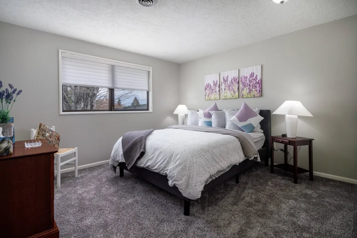 A serene bedroom with a queen bed adorned with decorative pillows, framed lavender artwork above the headboard, soft bedside lamps, and a wooden dresser. The room’s color palette and natural light evoke a peaceful, restorative mood—perfect for small families or friends.